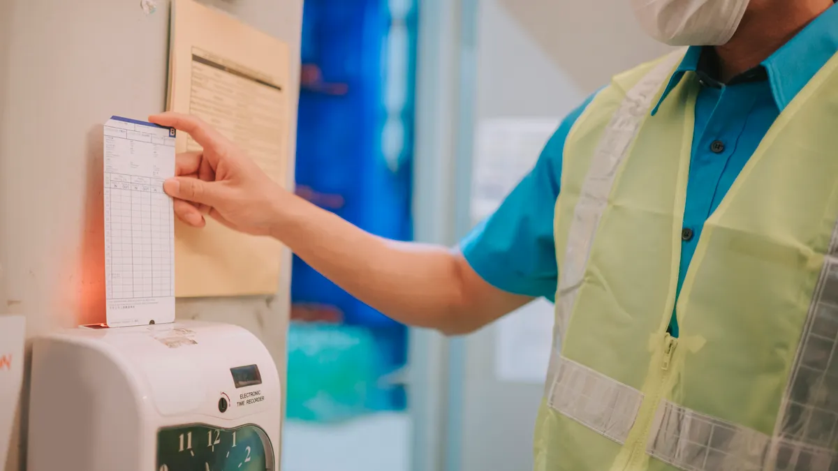 A worker in a safety vests uses a time card machine.