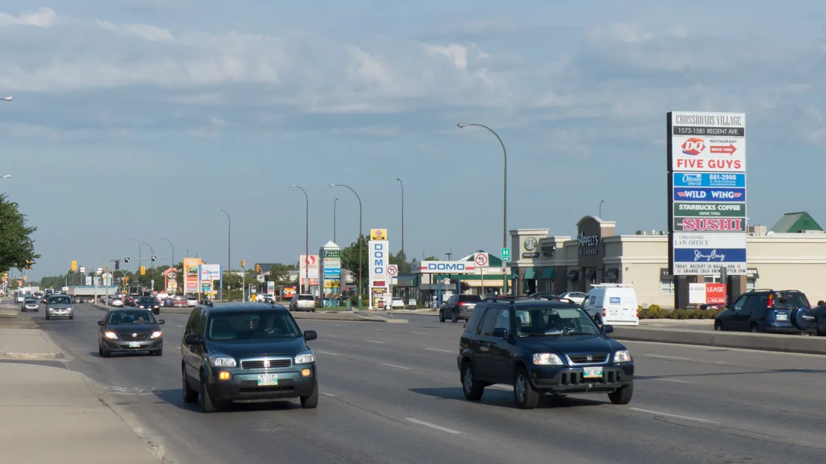 Cars driving on a highway lined with strip malls, and signs with names of retailers.