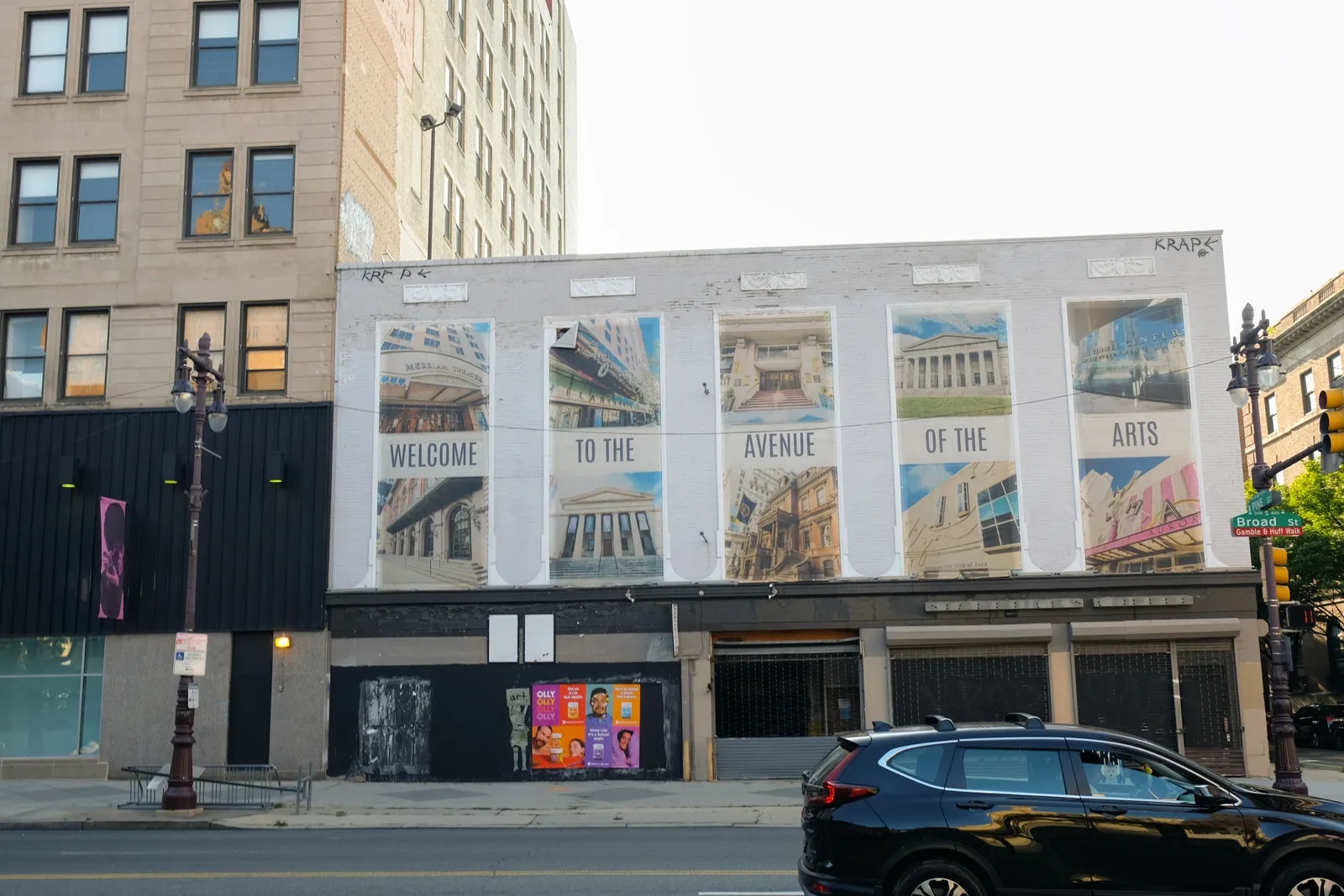 Alt text: Separated banners on a shuttered building read “Welcome to the Avenue of the Arts” as a car drives by.