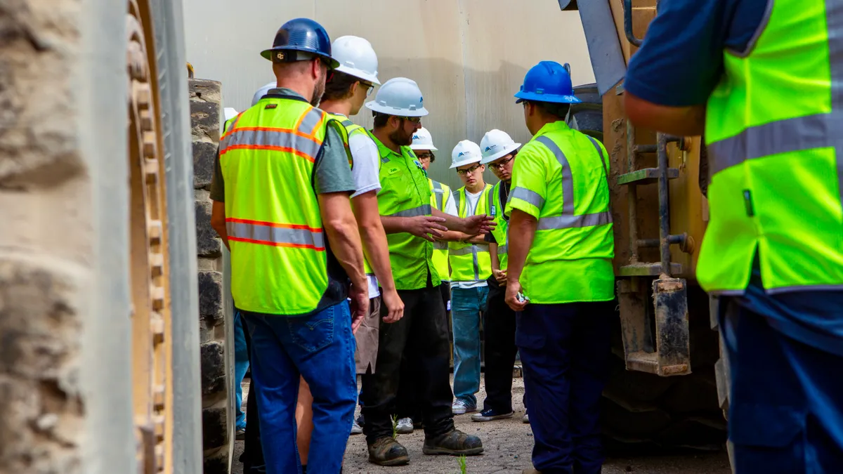 A group of people in hard hats and safety vests talk around heavy machinery.