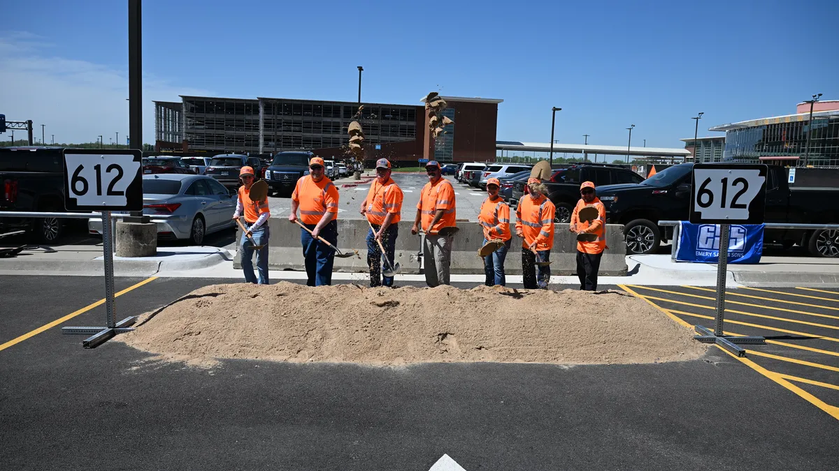 Seven people in orange reflective shirts and caps stand in a row, shoveling dirt from a pile between two 612 signs.