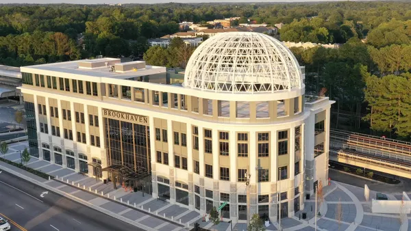 A large, newly constructed building looms above the treeline, with an open roof space and cars around.