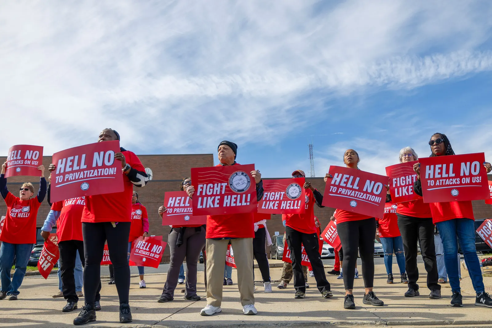 More than a dozen people wearing red shirts hold red signs that read “Hell No to Privatization”.
