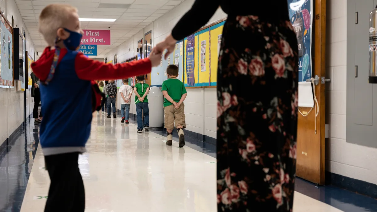 A young student wearing a face mask holds the hand of an adult in a school hallway as after young students walk down the hall.