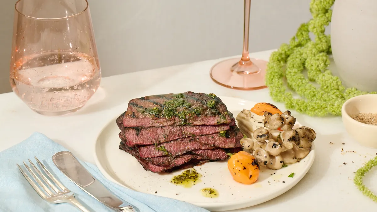A white plate with a stack of five grilled thin steaks with a green dressing on top. The plate also has mushrooms and small grilled peppers. The plate is on a table set for dinner.