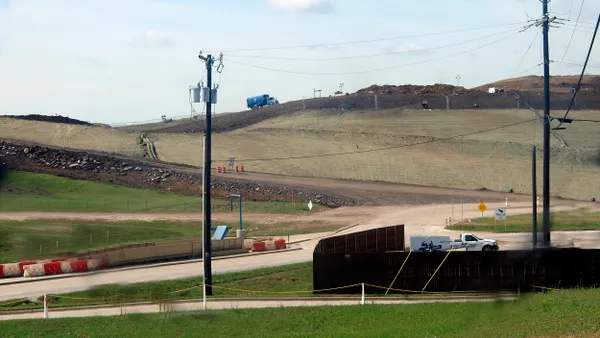 A wide view of a landfill site with grassy and dirt-covered hills, construction equipment, and a blue truck driving along a dirt road at the top of a slope. The foreground includes a paved access road with red-and-white barriers, utility poles with overhead wires, a small white pickup truck near a fenced area, and several warning signs along the road.