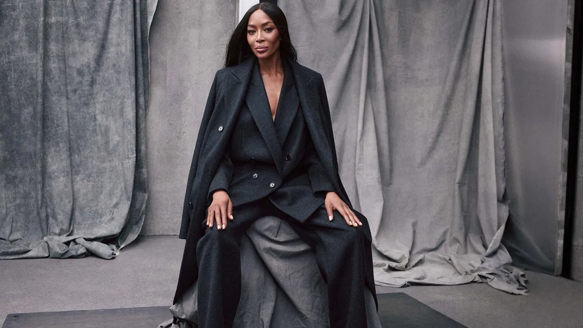 A model, Naomi Campbell, sits in front of a gray draped background wearing a dark gray flannel womenswear suit and coat, and stares at the camera with a slight smile.