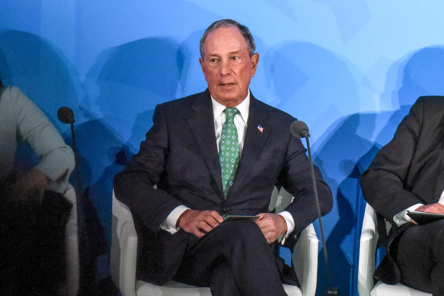 Michael Bloomberg is seated in the center of the frame wearing a black suit with an American flag pin on the left lapel, photo right, wearing a white shirt and green tie.