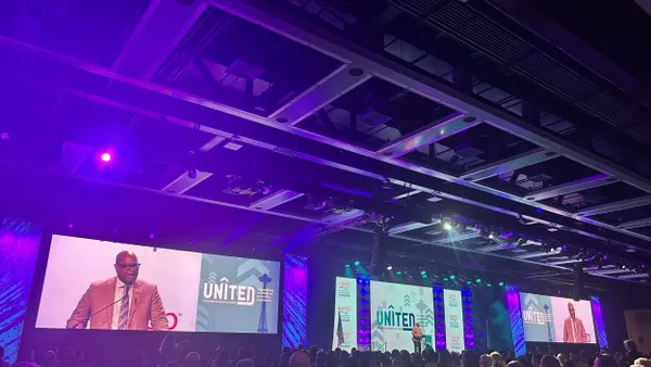 Three large screens appear above a stage that say "UNITED" with a person speaking who is displayed on the outer screens.