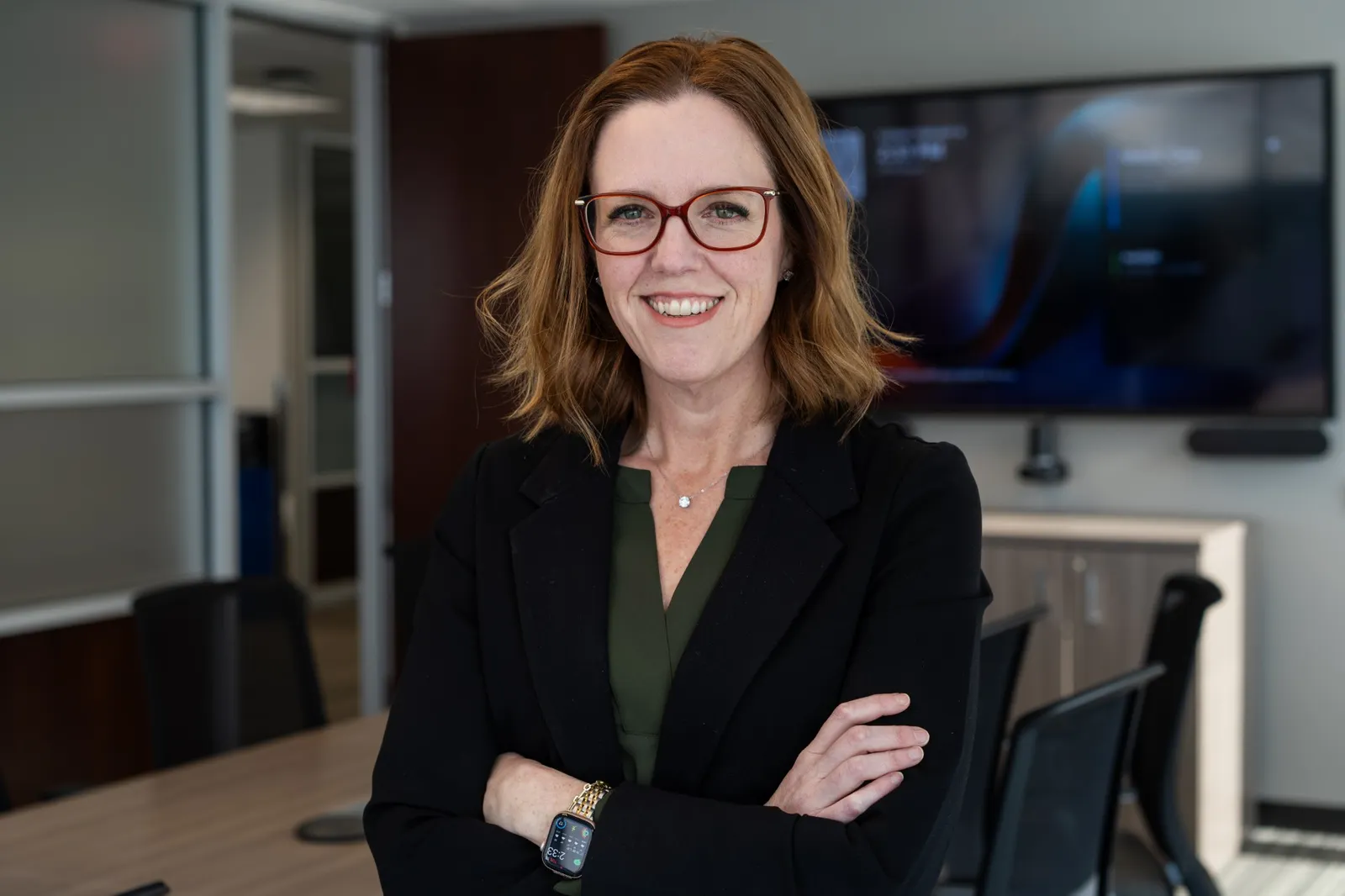 Headshot of a white woman, approximately 40-50, with light brown hair wearing red glasses, a green shirt and a black blazer