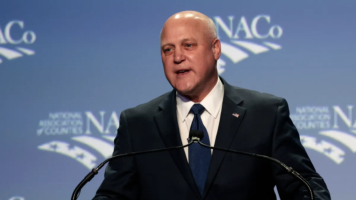 A man at a suit speaks at a podium with a blue backdrop.