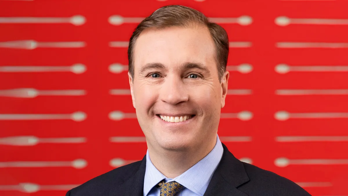 An image of a man in a suite smiling in front of a red backround with beigh spoons.