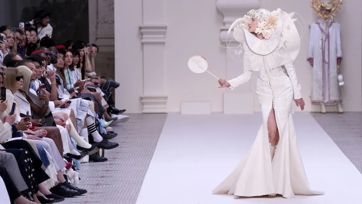 A modelon a runway wearing a in a cream gown and headdress leans toward the front row.