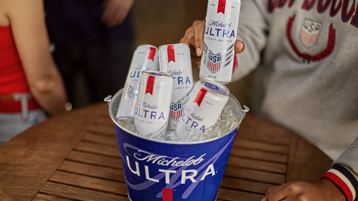 Cans of Michelob Ultra rest in a branded metal bucket atop a circular wooden table. A person in a USA soccer crewneck takes a bottle from the bucket.