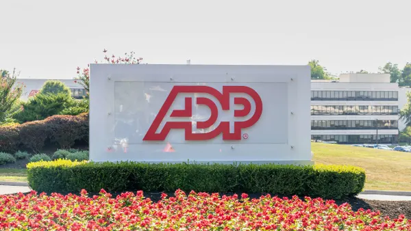 A white ADP sign with bold red letters, positioned in a landscaped area with vibrant red flowers and manicured bushes. The background shows the company’s headquarters building with rows of windows.