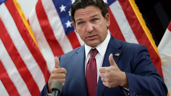 A man in front of American flags gives two thumbs up in front of a microphone.
