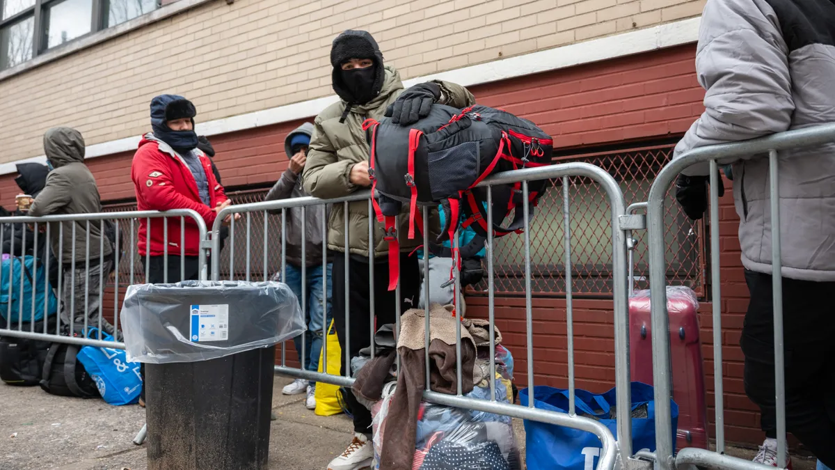 People in coats and masks stand in line against a building behind a metal fence. Many have large backpacks and bags.