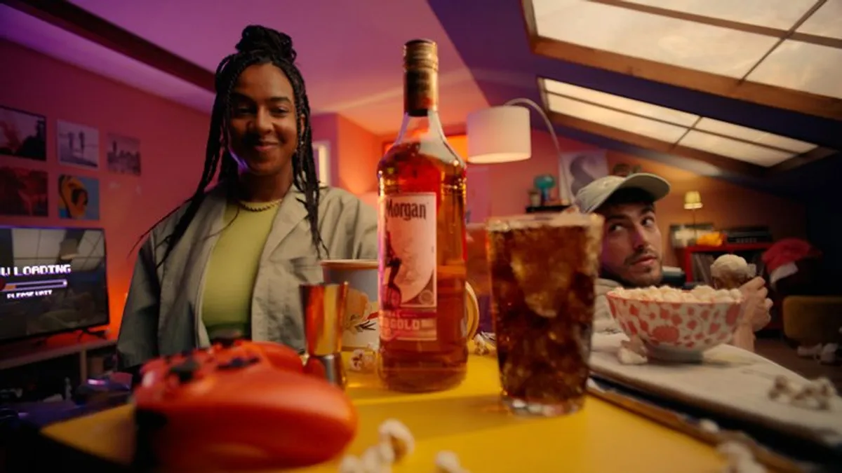 A bottle of Captain Morgan Rum sits on a table with a red video game controller as two people hang out.