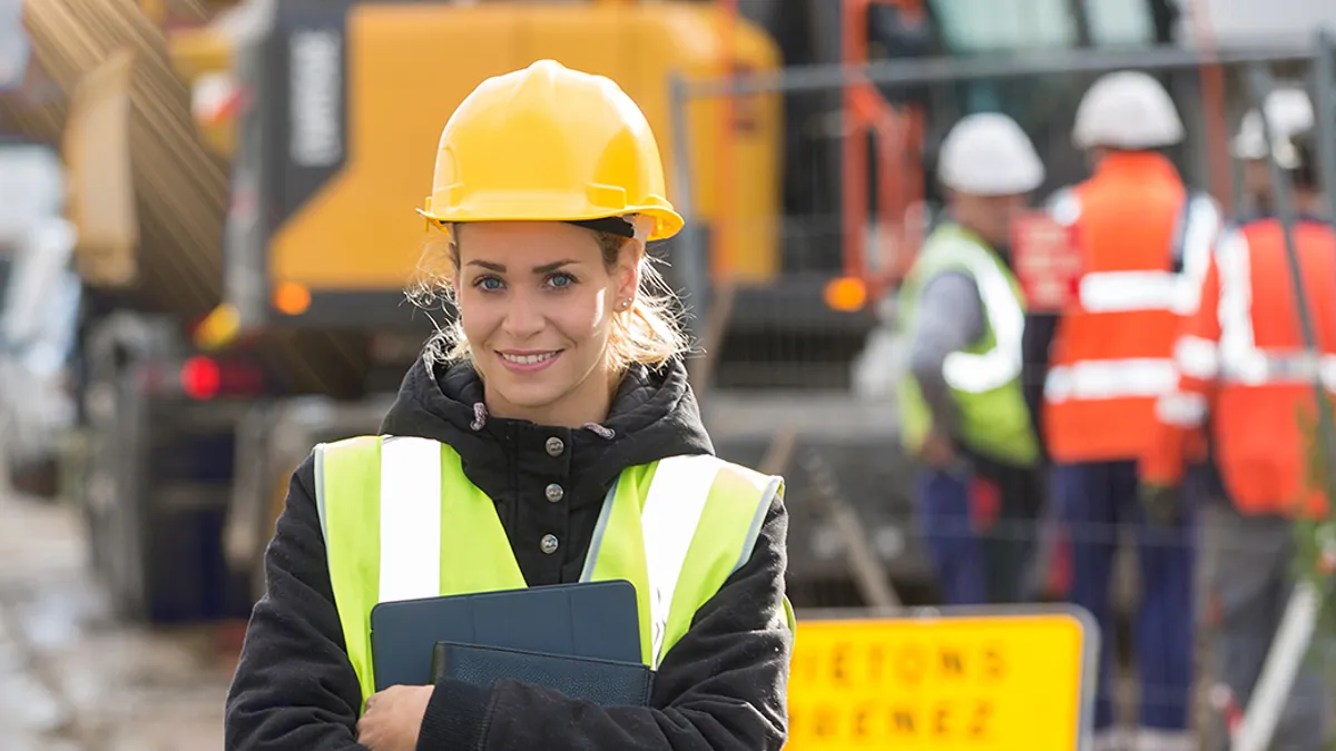 Woman in hard hat and construction vest posing in front of construction work site.