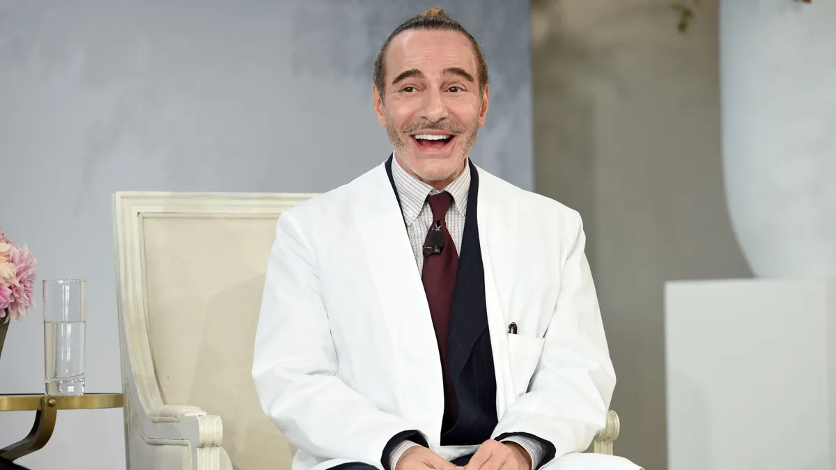 A man in a white suit smiles while sitting in a chair on stage.