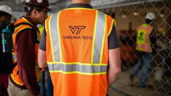 Two people stand with orange safety vests near a chain-link fence. One student has the logo of Virginia Tech on their back.