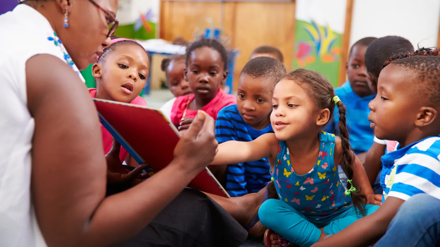 An adult sits on the floor holding a book with back toward camera. A group of young students sit facing the adult and look at the book.