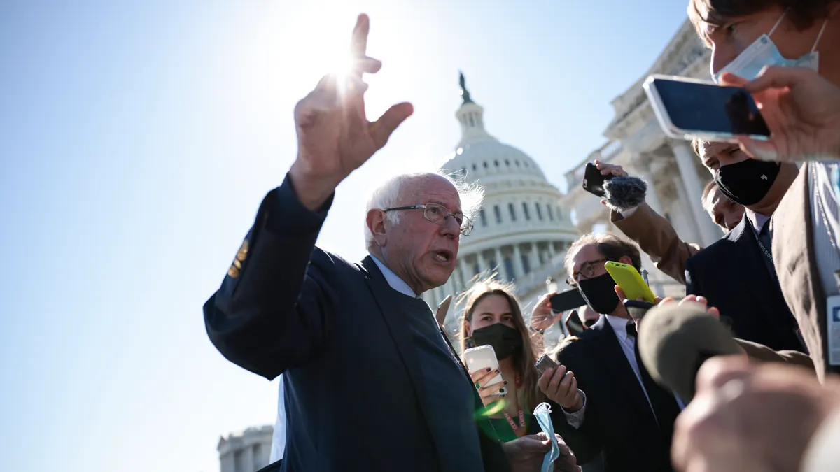 A picture from chest-up of Bernie Sanders speaking to reporters. The top of the US capitol building is visible behind him.