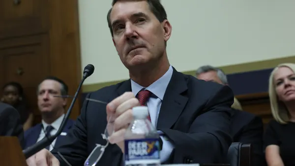 PNC CEO Bill Demchak holds his glasses and looks on during a Congressional hearing, as people sit behind him in the background.
