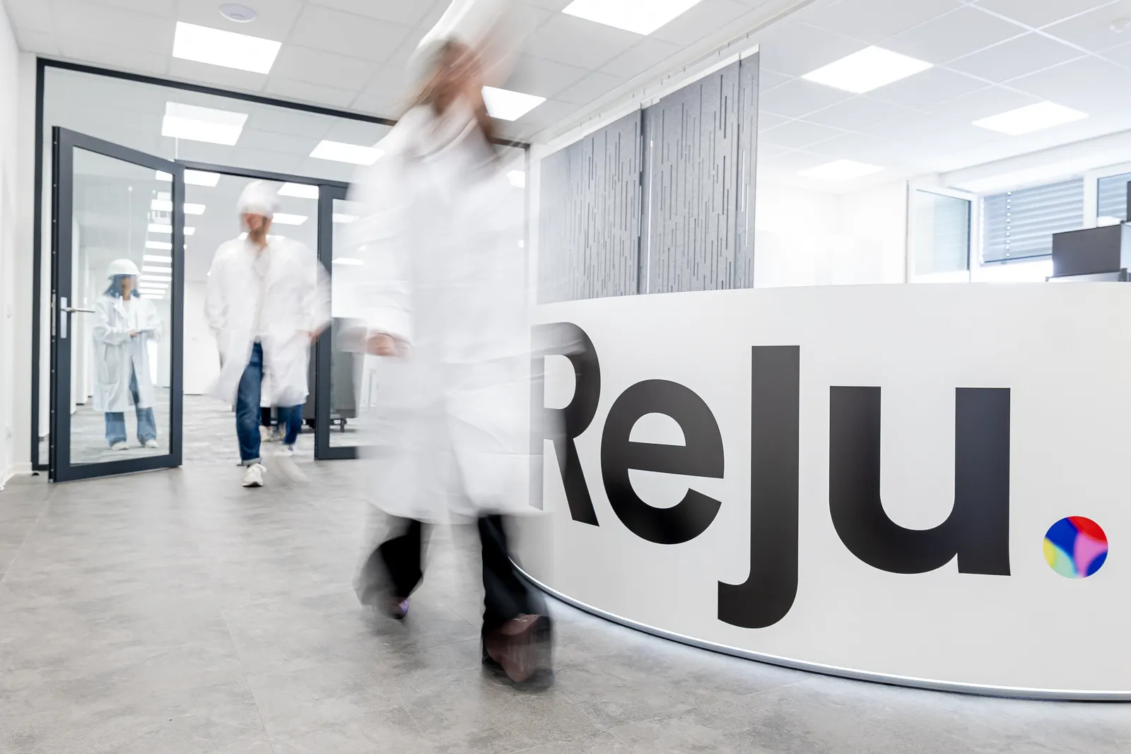 A time-lapse photo of workers walking by a Reju logo sign inside an office suite