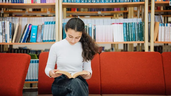 A student sits on a red couch in a library and looks down at a book in their lap.