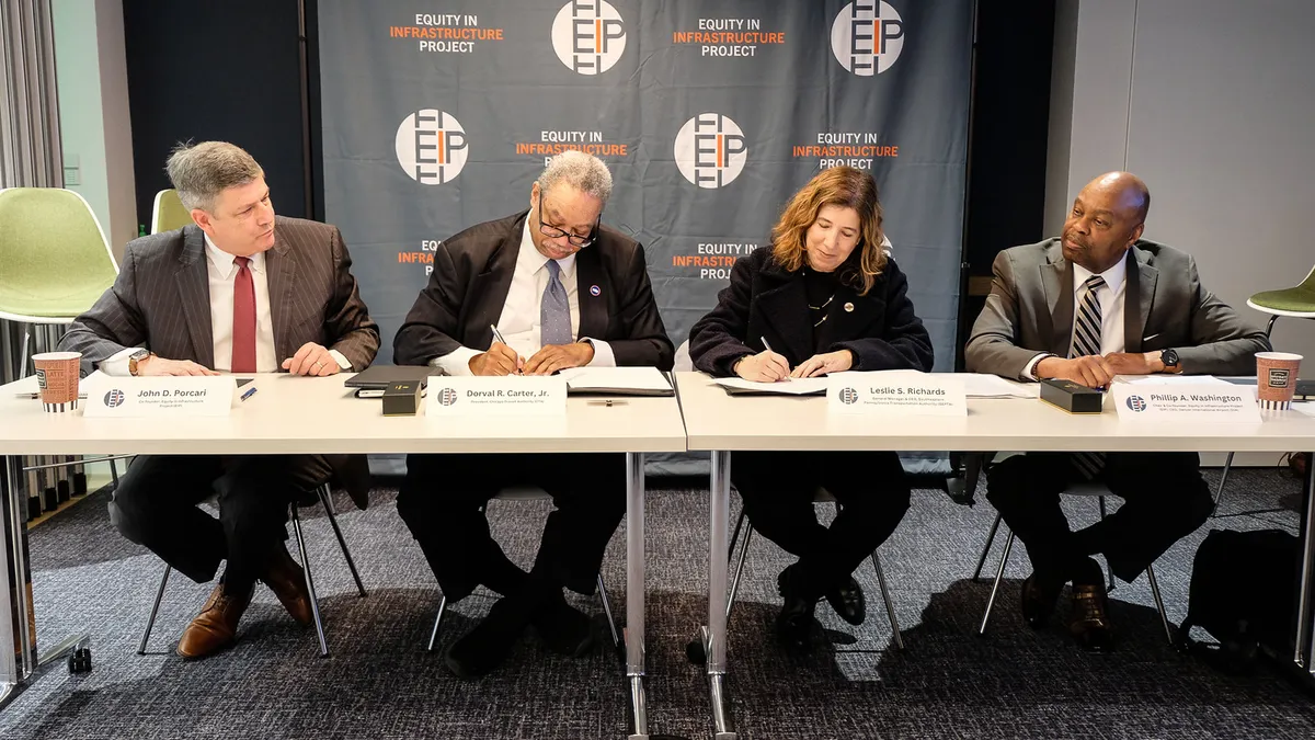 Four people in suits sit at a table, the two in the middle are signing documents.