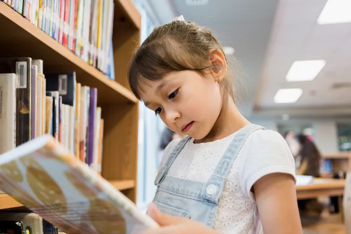 A student is looking down at an open book while standing near a book shelf