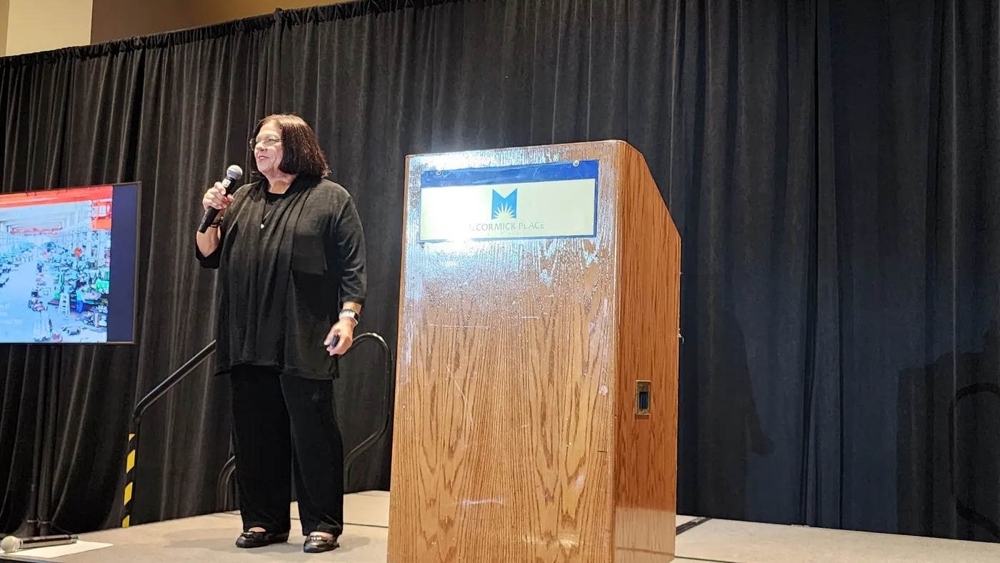 A person dressed in black and wearing glasses speaking into a microphone on stage, standing between a podium and a screen.