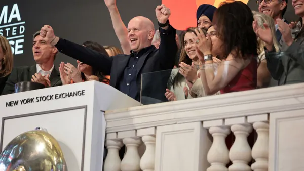 Mastercard CEO Michael Miebach stands in front of other people behind a podium at the New York Stock Exchange, with hands reaching out in celebration.