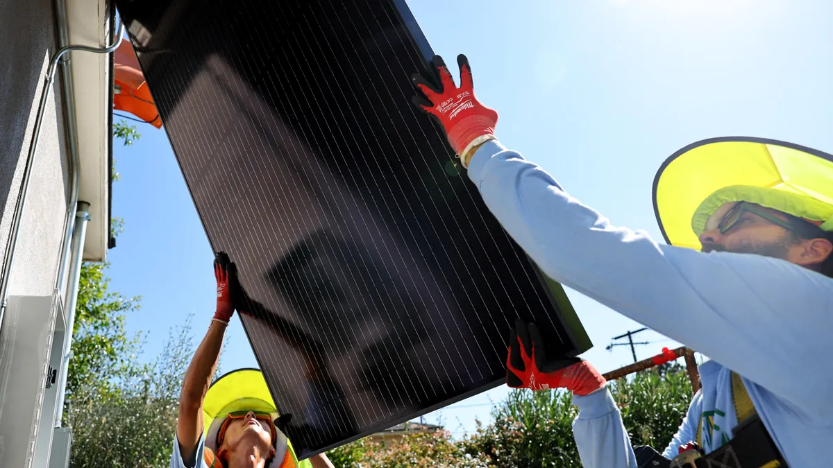 Two people wearing yellow wide-brimmed hats and orange work gloves hold a solar panel leaning on a home's roof.