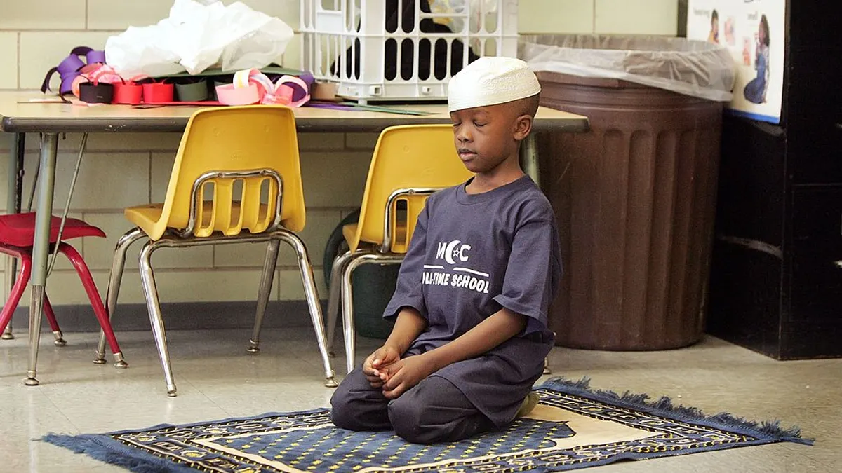 A boy bends in prayer on a prayer rug, wearing an Islamic head covering
