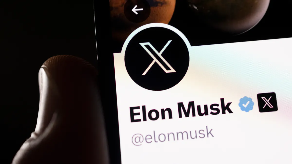 A hand holding up a smartphone displaying the X app profile of Elon Musk