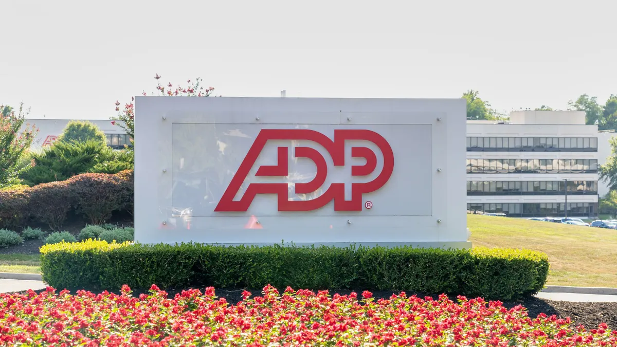 A white ADP sign with bold red letters, positioned in a landscaped area with vibrant red flowers and manicured bushes. The background shows the company’s headquarters building with rows of windows.