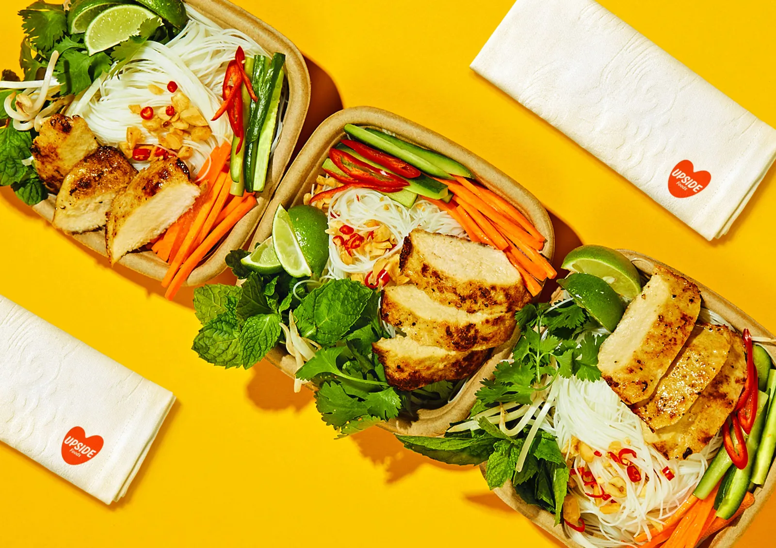 Three square paper containers of chicken chunks, greens and crudites with Upside Foods napkins nearby on a yellow background