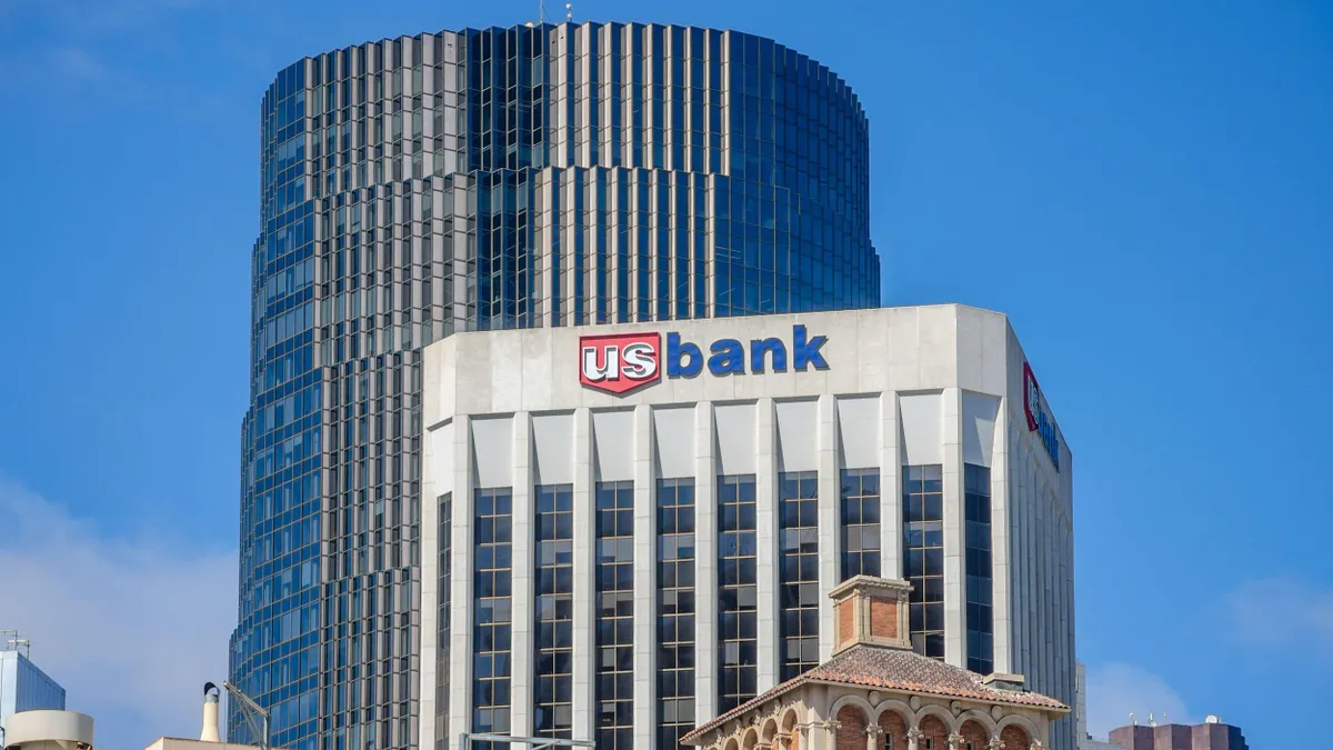 A tall modern office building with a white facade and the "US Bank" logo at the top, positioned in front of a taller cylindrical glass skyscraper.