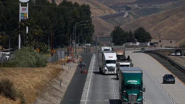 Trucks drive along an interstate in a line with a Shell gas station sign on the side of the road.