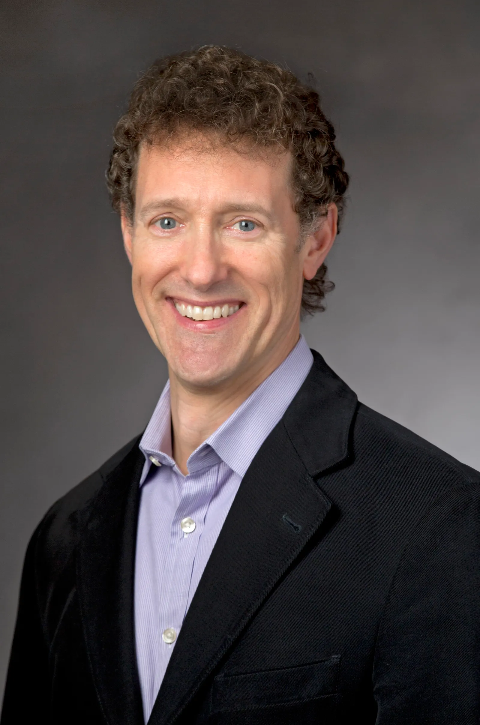 A headshot of Alex Cook wearing a black blazer against a dark backdrop.