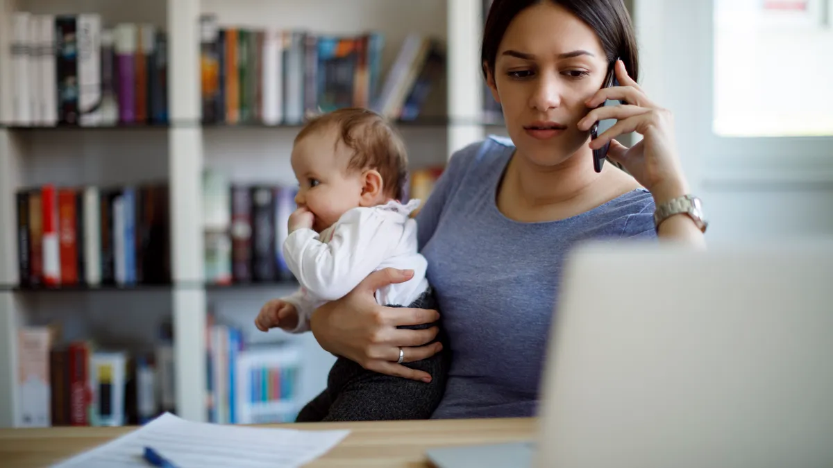 Adult sits at desk with an opening laptop and cell phone in one hand. In the other hand is an infant.