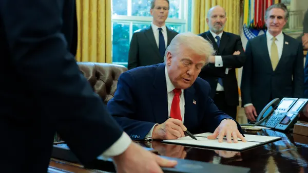 President Trump at his Oval Office desk signing an executive order surrounded by men.