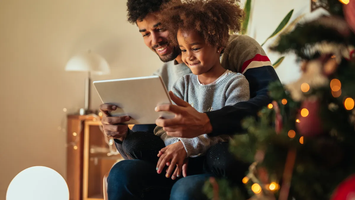 A man and young woman sit and look at a tablet next to a Christmas tree.