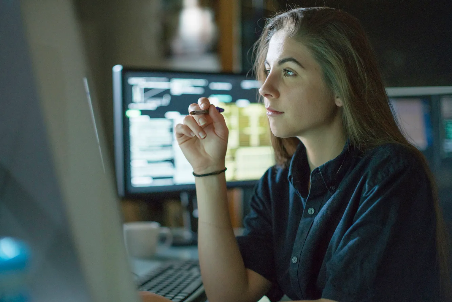 A young woman is seated at a desk surrounded by monitors displaying data, she is contemplating in this dark, moody office