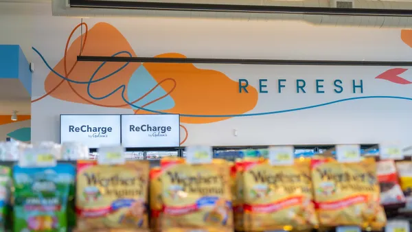 A photo of the wall inside of a store, seen over the top of several shelving units. Signs on the wall say "Refresh" and "Recharge by Gelson's."