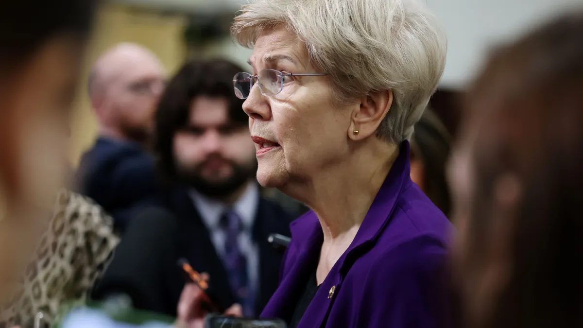 Close up of Senator Elizabeth Warren in a purple jacket speaking to reporters around her.