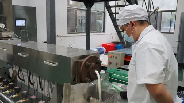 A person wearing a white shirt, cap and face mask stands next to industrial equipment that is producing a white material.