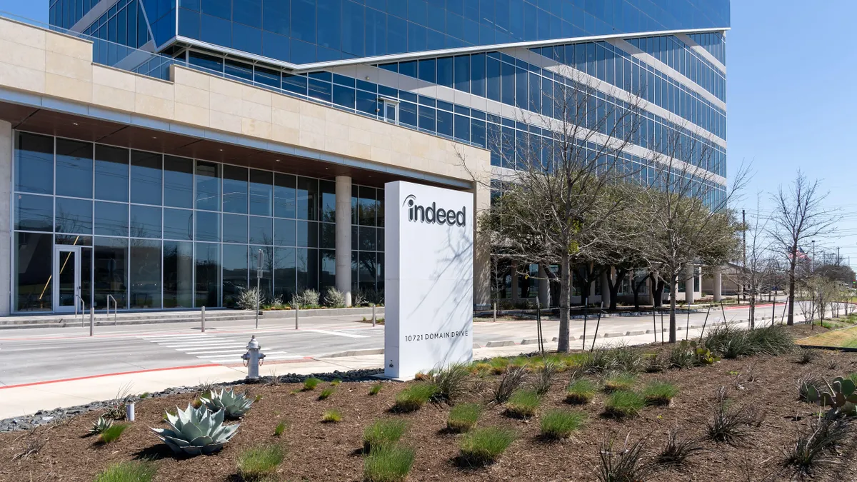 A modern glass office building with a large sign reading “indeed” on a white pedestal in front, surrounded by landscaped plants.